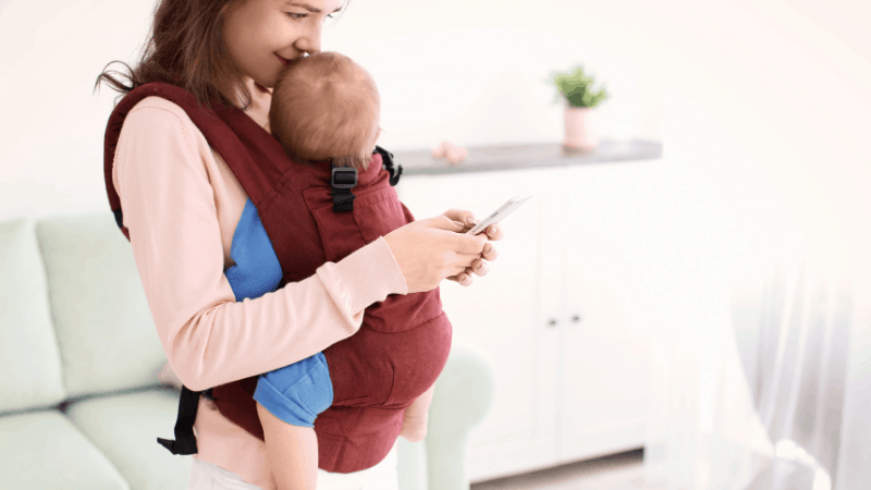 Mom holding baby in wrap looking down at phone.